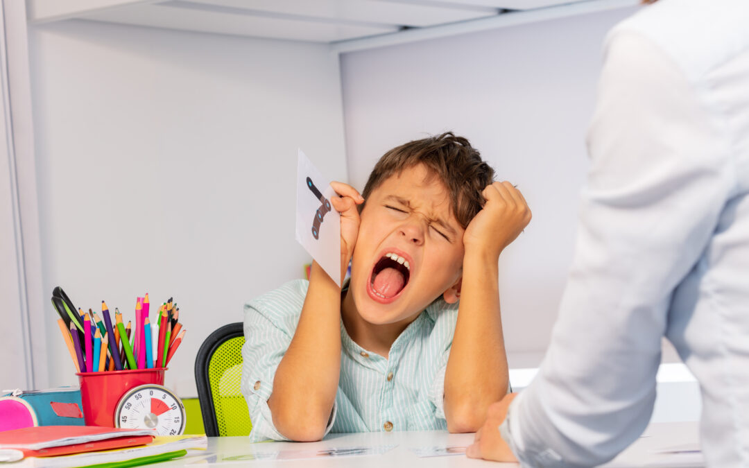 young boy is sitting at a desk with eyes closed, hands to his head, yelling and feeling frustrated. He is having trouble with emotional regulation while learning. An adult woman is sitting before him trying to do schoolwork with him.