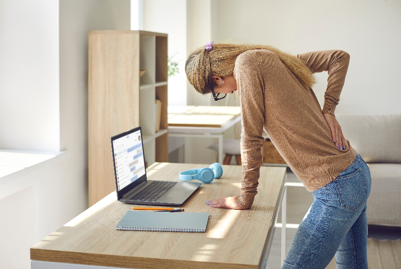 A blonde haired woman is standing up next to her desk. She has a laptop, appears to be working. She is looking down and holding her back. This woman suffers from chronic back pain which interferes with her life.
