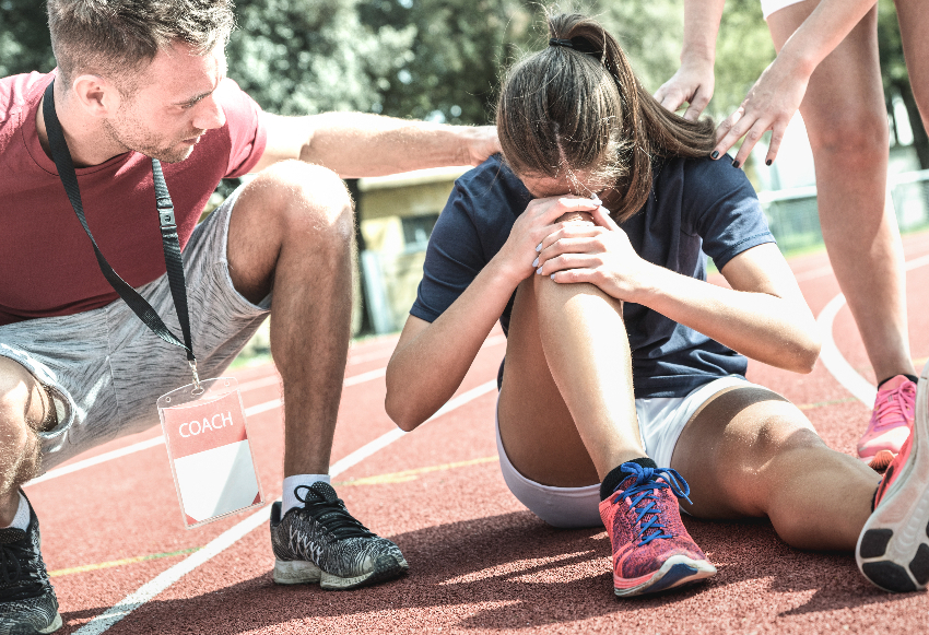 blond teen girl is sitting down on the high school track. Her head is to her knee, she is in pain. Her coaches are sitting around her. She just experienced a sports injury and will now need to navigate how to recovery from it.