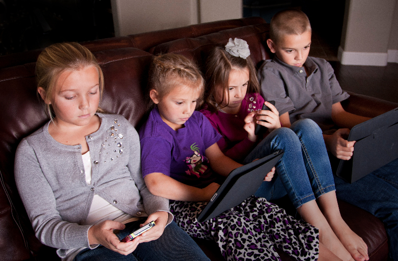 four kids sitting on a couch next to each other. Each one is having screen time, each on their own separate electronic device.